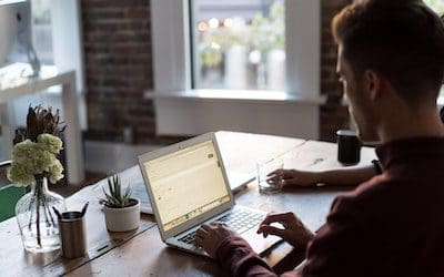 male typing at a laptop on a wooden desk inside a well decorated brick community building near a window