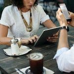 Image of woman using a tablet and other person using a handheld device while sitting at a table having coffee